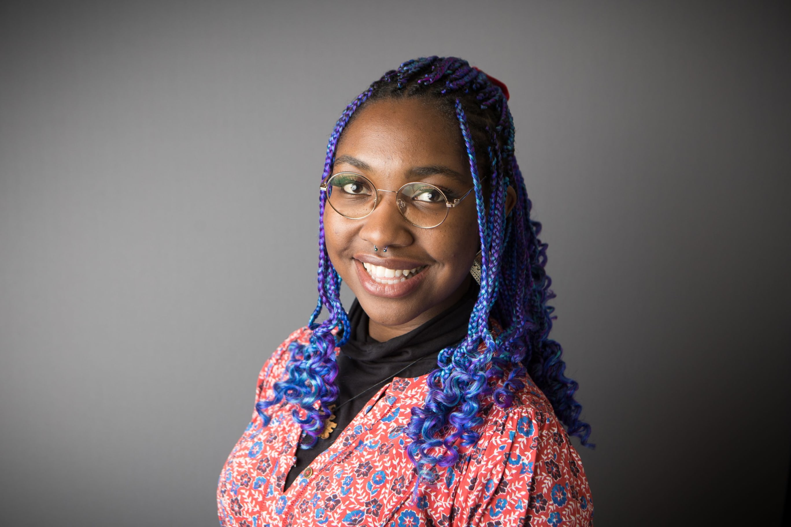Headshot of Millicent, a black woman with long blue and purple curly braids, glasses and a patterned top